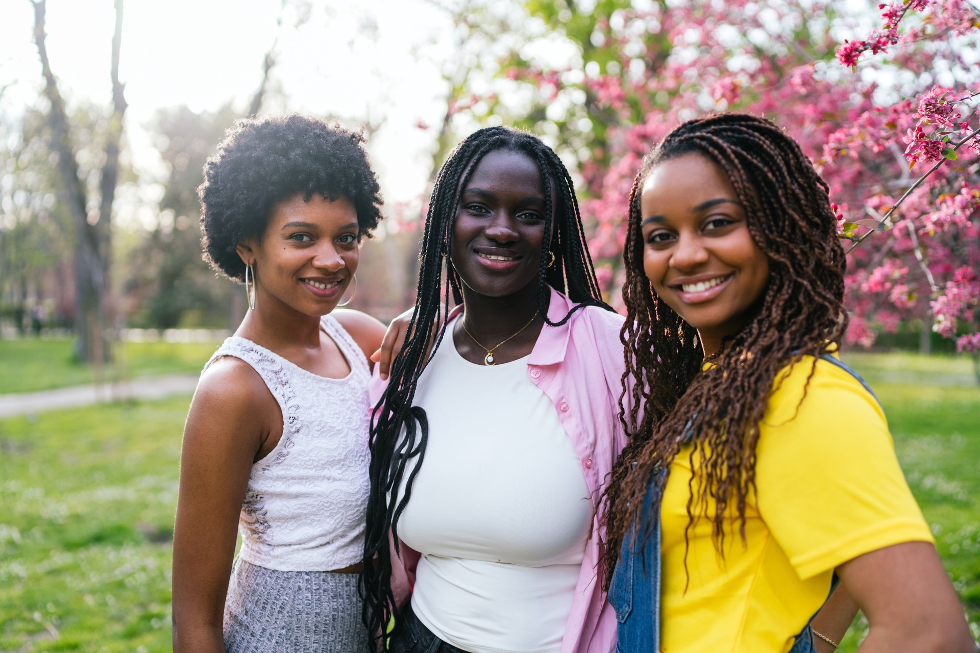 Three cheerful African women posing in a park surrounded by pink blossoms.